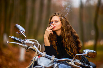 Fototapeta na wymiar A beautiful long-haired woman smoking on a chopper motorcycle in autumn landscape on a forest road