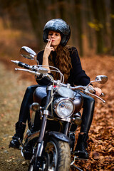 Obraz na płótnie Canvas A beautiful long-haired woman smoking on a chopper motorcycle in autumn landscape on a forest road
