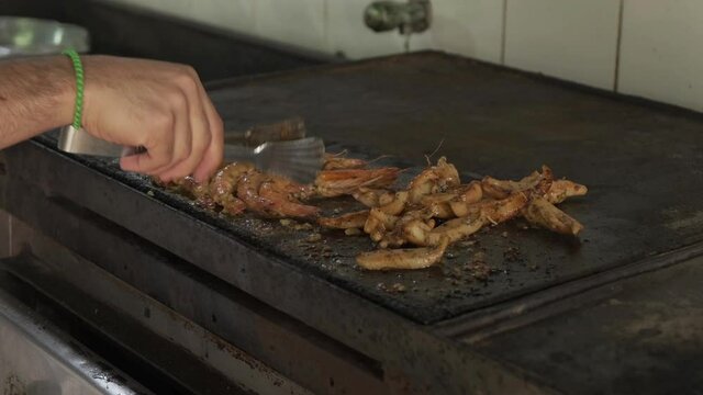 Chef prepares fish and seefood at the city market. Shrimps are fried on the grill, he puts it in a row. Close-up view