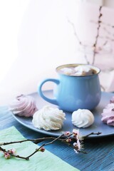 Cup of coffee with meringue, flowering cherry branches in water in a glass jar, sweet dessert on a plate on a wooden windowsill, spring season, home comfort