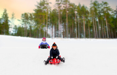 childhood, sledging and season concept - happy little children sliding on sleds down snow hill in winter over snowy forest or park background