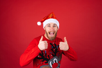 Red-bearded man in a santa hat and a christmas sweater on a red background cheerfully smiles thumbs up