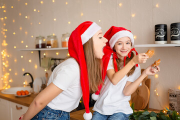Fototapeta premium Good Christmas morning. Sisters eat cookies at the kitchen table. The girls are wearing Santa Claus hats. A time of miracles and fulfillment of desires. Merry Christmas.