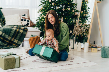 Obraz na płótnie Canvas Young Caucasian diverse redhead mommy sitting on the floor with toddler unpacks Christmas present. New Year concept. Family traditions of gift giving. Decorated living room and cozy interior.