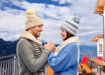 Fototapeta premium travel, tourism and winter holidays concept - happy romantic couple in knitted hats and scarves holding hands over mountains and ski resort background