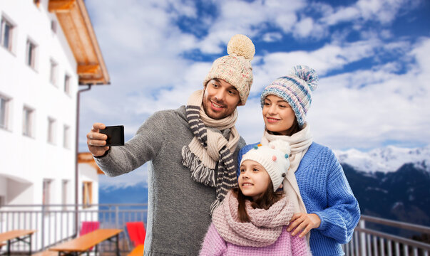 travel, family and winter holidays concept - happy mother, father and little daughter in winter clothes taking selfie by smartphone over mountains and ski resort background