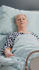 Portrait of sick patient laying in hospital ward bed, looking at camera. Senior woman with nasal oxygen tube and IV drip bag resting to cure diagnosis for healthcare and recovery.