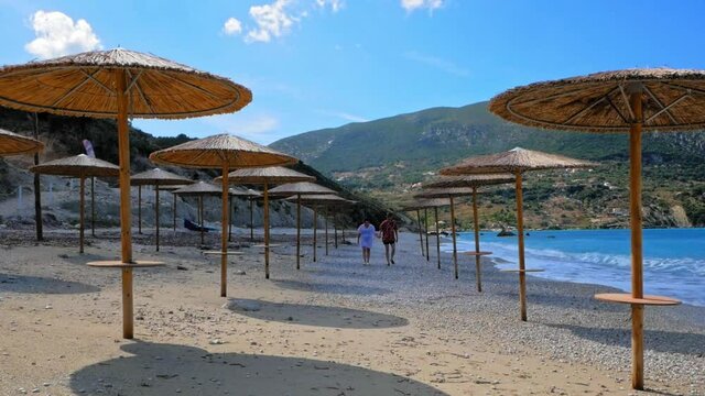 Happy Couple Holding Hands, Walking Together On The Shore With Native Beach Umbrellas In Agia Kiriaki Beach, Zola In Kefalonia, Greece. - Long Shot, Slow Motion