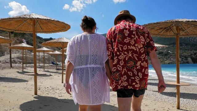 Agia Kiriaki Beach, Zola, Kefalonia, Greece - Back View Of Romantic Couple Holding Hands While Walking At Summer Beach With Native Umbrellas And Sea View Background. - Long Shot, Slow Motion