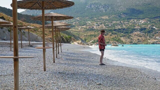 Man Wearing Hat And Summer Beach Floral Outfit Throwing Pebbles Into The Sea In Agia Kiriaki Beach, Zola, Kefalonia, Greece. - Medium Shot