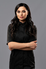 Front view of beautiful caucasian woman in black medical uniform standing in studio. Competent female cosmetologist holding arms crossed over grey background.