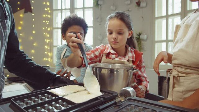 Cute little girl pouring batter in waffle maker and passing ladle to friend while learning cooking with chef on culinary masterclass