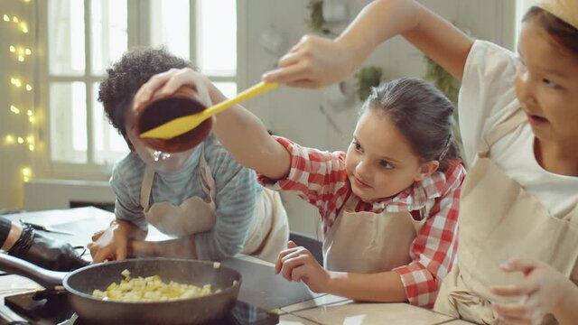 Group of cute multiethnic kids adding honey to diced fruit and stirring ingredients in pan while cooking together on culinary class