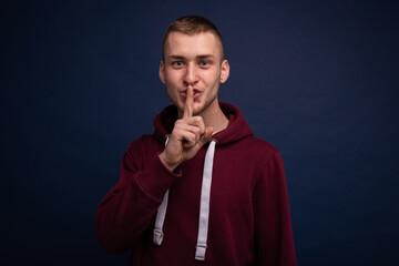Young man in red hoodie and jeans poses on a blue background and shows quietly
