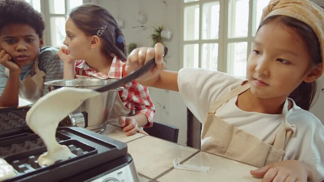 Little Asian girl pouring batter in waffle maker learning cooking with friends and chef on culinary masterclass