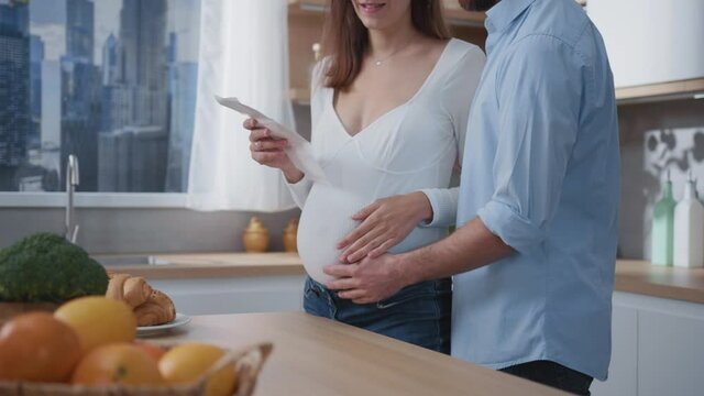 Young couple of future parents pregant wife with husband expecting child watching an ultrasound echography photograph in modern stylish cozy home kitchen.