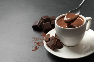 Spoon with yummy hot chocolate above cup on dark table, space for text