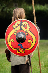 Back of Viking man standing with spear in hand showing red wooden shield with yellow pattern