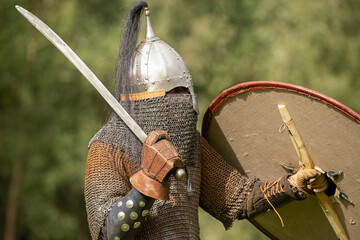 medieval soldier knight in armour chain mail coif holding sword and shield and wearing helmet and leather glove against green background during re-enactment