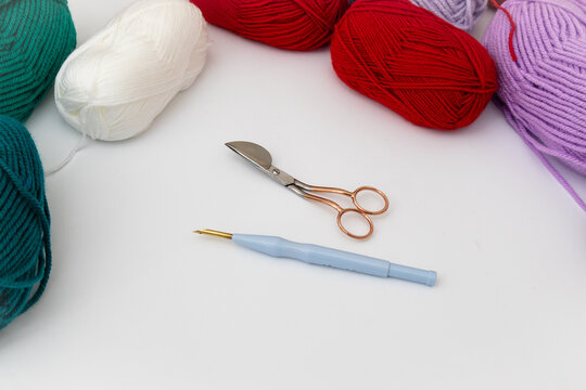 Rounded scissors, punch needle, and skeins of yarn in different colors: white, red, green and purple on white background