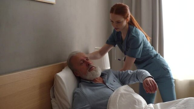 Caring young female home caregiver helping senior adult male patient to lying on bed in bedroom. Professional nurse helping old man to lying on bed in luxury hospital room. Shooting in slow motion.