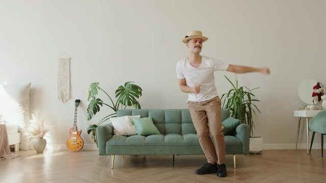 Funny modern fashion hipster with mustache wearing hat, round pink sunglasses dancing, fooling around, lying on sofa in modern loft apartment. Millennial generation killing time lifestyle