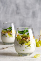 Two glasses of plain yoghurt with pieces of fresh kiwi fruits, white grapes, granola, and mint leaves on an old piece of wood with a grey background. 