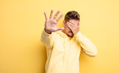 young handsome man covering face with hand and putting other hand up front to stop camera, refusing...