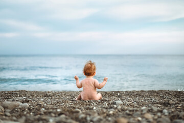 Fototapeta na wymiar A baby, a tiny boy is sitting with his back alone on the seashore, a beach, pebbles, the child's face is not visible.