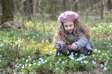 Picture of a girl in a beret and coat sitting on her haunches in the grass and flowers