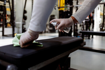 Young man cleaning and disinfecting with sanitizer the gym equipment. Hands of men at the modern gym using the cloth and disinfection liquid in spray bottle to clean the exercising machines in the gym