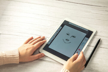 Young creative designer holding stylus pen drawing on screen of digital tablet on wooden desk with copy space, modern digital art top view