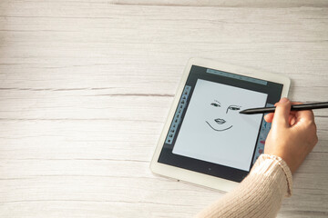 Young creative designer holding stylus pen drawing on screen of digital tablet on wooden desk with copy space, modern digital art top view