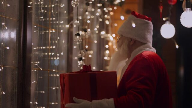A real Santa Claus with glasses and a beard on the outdoor comes to the window of the house with a gift on the night before Christmas