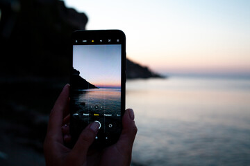 woman hands holding mobile phone at sunrise over sea bay. Taking photo of sunset over rocks in...