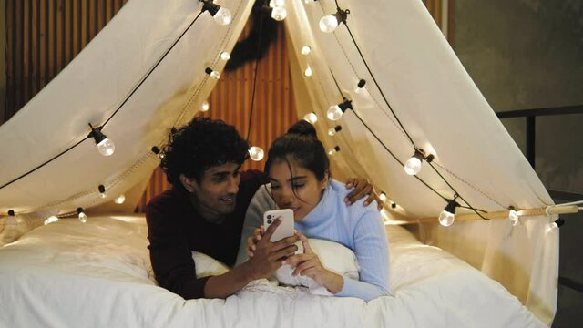 Cute young indian couple students lying in cozy place bed tent with lights on christmas, playing games on mobile phone, making purchase for new year on christmas night, having insomnia, love story