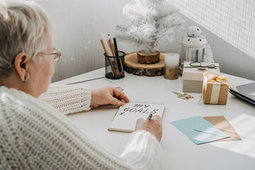 Fototapeta na wymiar Healthy New Years resolutions for older adults. Senior mature old woman in white sweater writing handwritten text My Goals in open notepad on the table. Planning and setting goals