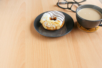 Donut with cream and nuts toppings on the plate, a cup of coffee and eyeglasses on the wooden table 