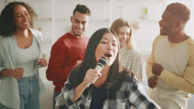 Happy party at home. Young positive asian woman singing song with microphone, group of cheerful diverse friends dancing