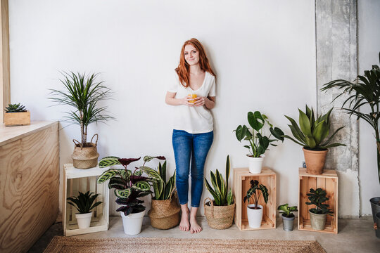 Smiling woman with glass of juice standing amidst houseplants at home