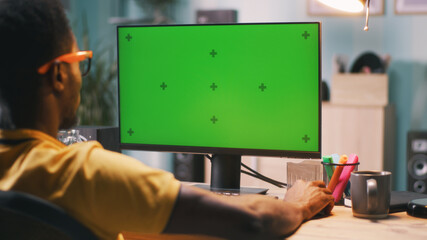 African American guy in glasses sitting at table and browsing computer with chromakey screen during work in home office