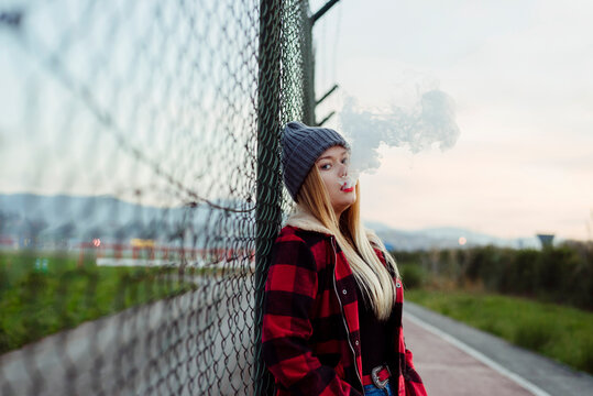 Young woman with wool hat leaning on the fence and smoking