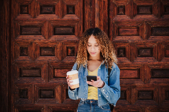 Curly haired woman holding reusable cup using smart phone by wooden door