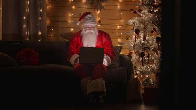  General plan Front view A real Santa Claus is working with a laptop at night with glasses in the light of Christmas lights on the background of a Christmas tree. Remote work