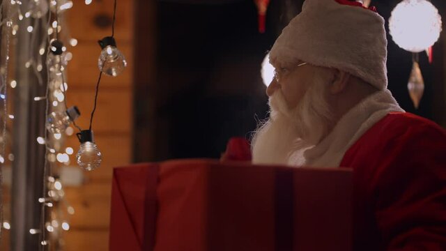 Portrait of a real Santa Claus with glasses and a beard on the outdoor comes to the window of the house with a gift on the night before Christmas and looks inside. Santa brings a gift