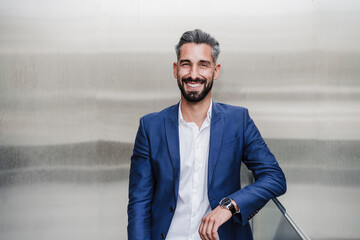 Mid adult businessman smiling while leaning on railing in front of silver colored wall