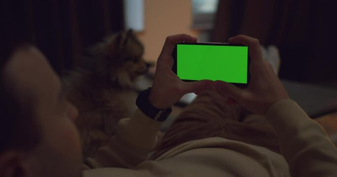 Man Using Smartphone in Horizontal Mode with Green Mock-up Screen. Resting at Home with small dog looking at Phone Screen Watching Film, Video Content. Using Mobile Apps. Chroma Key. Device.