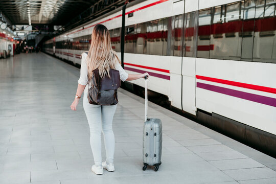 Young female passenger wearing backpack standing with wheeled luggage at railroad station platform