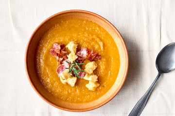 Pumpkin cream soup with roasted bacon and croutons in clay plate on the table with linen tablecloth. Autumn vegetable creamy soup puree. Healthy seasonal eating.