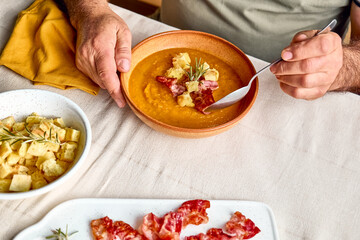Man eating pumpkin cream soup with roasted bacon and croutons in clay plate on the table with linen tablecloth. Autumn vegetable creamy soup puree. Healthy seasonal eating.